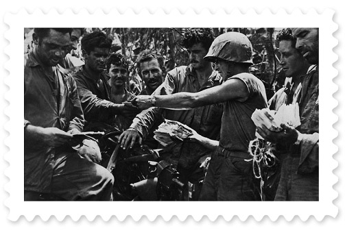 A black-and-white photograph of a soldier handing out mail to a group of fellow soldiers.