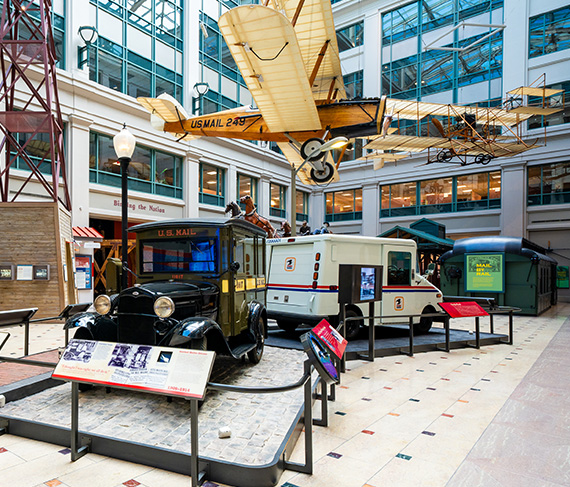 A colorful photograph of an older and newer version of a mail truck on display in a brightly lit atrium inside the National Postal Museum.